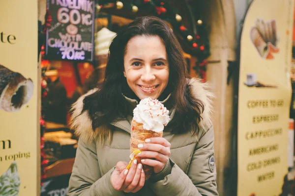 A beautiful girl in a warm jacket eats trdelnik or Trdlo with cream in her hands, in the winter in the Czech Republic, Prague at the Christmas market — Stock Photo, Image