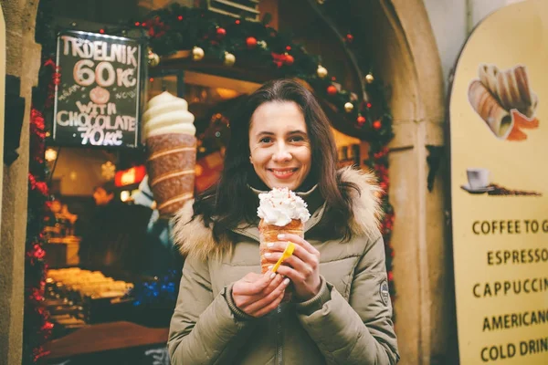Krásná dívka v teplé bundě jí trdelník nebo Trdlo se smetanou v dlaních, v zimě v České republice, Praha na vánočním trhu — Stock fotografie