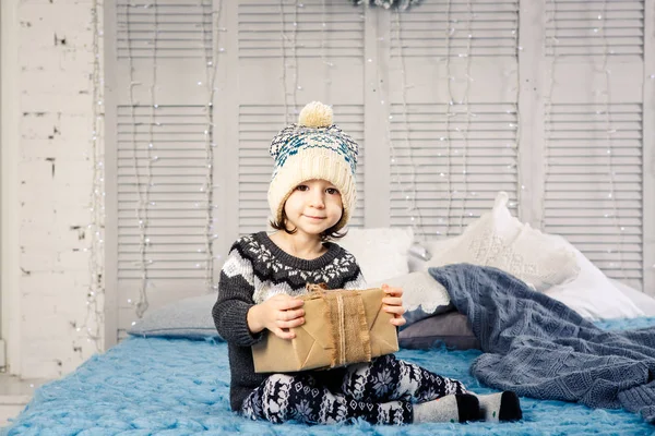 Niña el niño sentado en pijama y sombrero en la cama con guirnalda de bombillas con cajas de regalos envueltos en un papel no coloreado decorado con conos en azul de punto coverlet.Christmas concepto — Foto de Stock