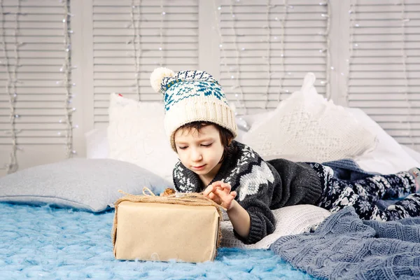 Kleines Mädchen das Kind in Pyjama und Hut auf dem Bett mit Girlanden von Glühbirnen mit Geschenkschachteln in einem farblosen Papier mit Zapfen auf blauem Strickbezug verziert sitzen.Weihnachtskonzept — Stockfoto