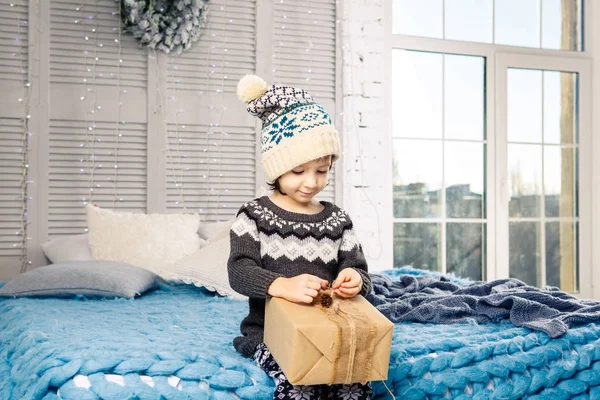 Niña el niño sentado en pijama y sombrero en la cama con guirnalda de bombillas con cajas de regalos envueltos en un papel no coloreado decorado con conos en azul de punto coverlet.Christmas concepto — Foto de Stock