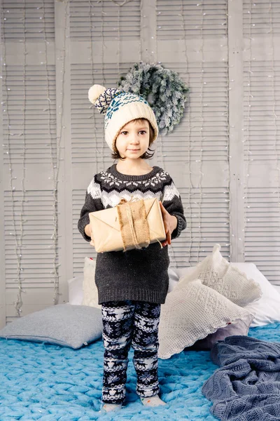 Μικρό κορίτσι το παιδί κάθεται στο πιτζάμες και καπέλο στο κρεβάτι με γιρλάντα από λάμπες φωτός με δώρα κουτιά τυλιγμένο σε ένα μη-χρωματισμένο χαρτί διακοσμημένα με κώνους για μπλε πλεκτό κουβέρτα. Έννοια των Χριστουγέννων — Φωτογραφία Αρχείου