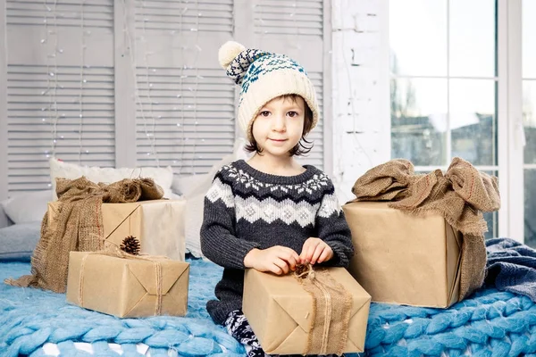 Niña el niño sentado en pijama y sombrero en la cama con guirnalda de bombillas con cajas de regalos envueltos en un papel no coloreado decorado con conos en azul de punto coverlet.Christmas concepto — Foto de Stock