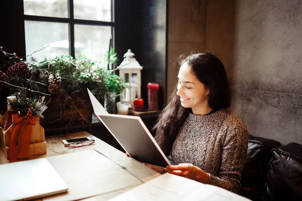 Zakelijke meisje bestuderen van het menu in restaurant ingericht met Kerstmis décor.sits in de buurt van het venster op bewolkt winterdag aan houten tafel. Gekleed in warm grijze trui. Op de tafel, de telefoon en de bril — Stockfoto