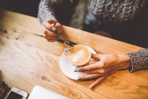 Vackra business tjej dricka kaffe ur vit kopp i restaurangen dekorerad med jul décor.sitting nära fönster på ett träbord. Klädd i varm grå tröja. På bordet, telefon och glasögon — Stockfoto