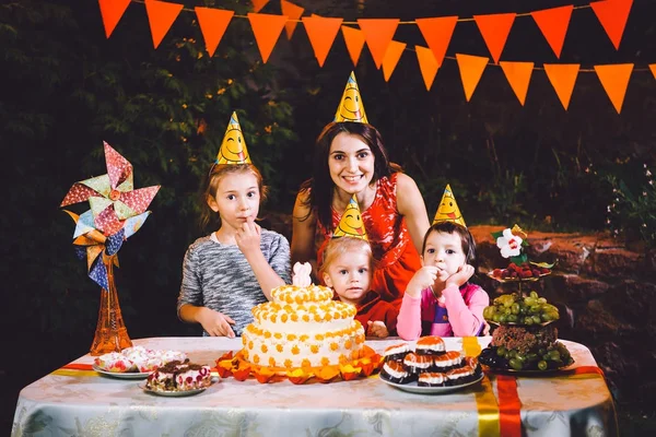 En stor mor och tre barn på en festlig bord med godis och en tårta i gården inredda med ljus och en krans av lampor. Begreppet en familj barns födelsedagsfest — Stockfoto