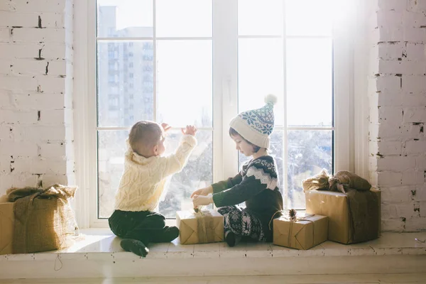 Los niños hermanos y hermanas de edad preescolar se sientan junto a la ventana en un soleado día de Navidad y juegan con cajas de regalos envueltas en papel. Están vestidos con ropa de lana caliente de punto y sombrero. — Foto de Stock