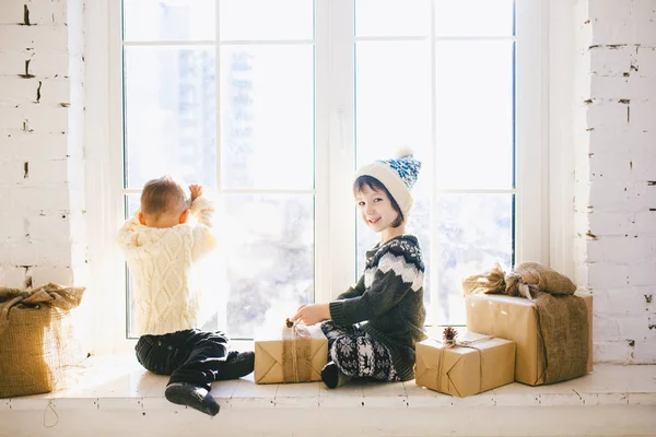 Los niños hermanos y hermanas de edad preescolar se sientan junto a la ventana en un soleado día de Navidad y juegan con cajas de regalos envueltas en papel. Están vestidos con ropa de lana caliente de punto y sombrero. — Foto de Stock