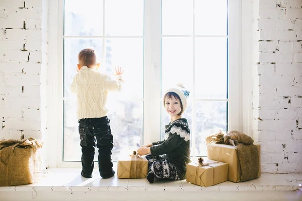 Τα παιδιά του αδελφού και της αδελφής της προσχολικής ηλικίας να καθίσει δίπλα σε παράθυρο σε μια ηλιόλουστη ημέρα των Χριστουγέννων και παίζουν με τα κουτιά δώρων τυλιγμένο σε χαρτί. Είναι ντυμένος πλεκτό ζεστά Μάλλινα ρούχα και καπέλο. Μέσα στο σπίτι — Φωτογραφία Αρχείου