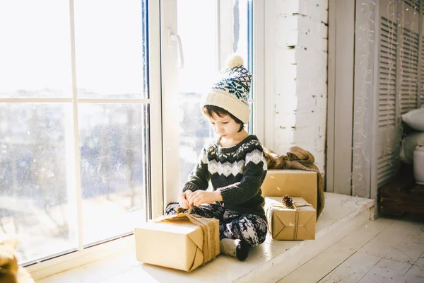 Niño está sentado junto a la ventana en un día soleado de Navidad y se besa con regalos en cajas envueltas en papel.Vestido con ropa de lana caliente de punto y un sombrero. Dentro de la casa con un elegante interior — Foto de Stock