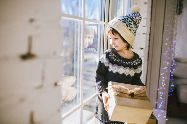 Gyermek ül az ablaknál egy napsütéses napon a karácsony, és teszi az ajándékok a doboz fóliába csomagolva, paper.dressed kötött meleg gyapjú ruhát és kalapot. A házban stílusos belsőterekkel — Stock Fotó