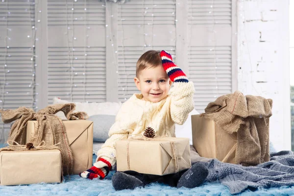 Pequeño niño divertido juguetón un niño se sienta en una cama en el día de Navidad con cajas de regalo en suéter de punto de lana blanca y grandes manoplas brillantes en él y se ríe en voz alta. En el interior hay una decoración festiva — Foto de Stock