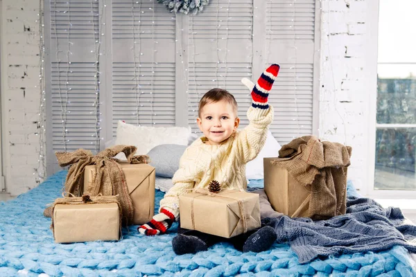 Pequeño niño divertido juguetón un niño se sienta en una cama en el día de Navidad con cajas de regalo en suéter de punto de lana blanca y grandes manoplas brillantes en él y se ríe en voz alta. En el interior hay una decoración festiva — Foto de Stock