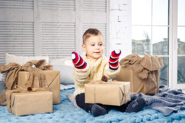 Pequeño niño divertido juguetón un niño se sienta en una cama en el día de Navidad con cajas de regalo en suéter de punto de lana blanca y grandes manoplas brillantes en él y se ríe en voz alta. En el interior hay una decoración festiva — Foto de Stock