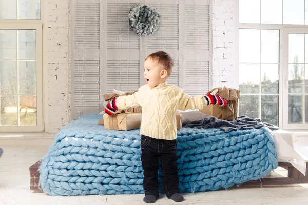 Pequeño niño divertido juguetón un niño se sienta en una cama en el día de Navidad con cajas de regalo en suéter de punto de lana blanca y grandes manoplas brillantes en él y se ríe en voz alta. En el interior hay una decoración festiva — Foto de Stock