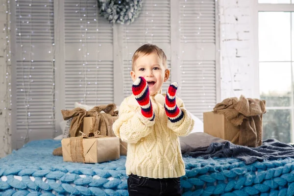 Pequeño niño divertido juguetón un niño se sienta en una cama en el día de Navidad con cajas de regalo en suéter de punto de lana blanca y grandes manoplas brillantes en él y se ríe en voz alta. En el interior hay una decoración festiva — Foto de Stock