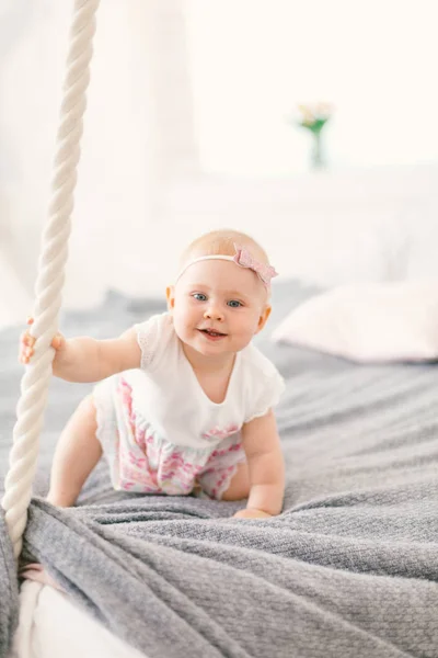 Kleines Mädchen, ein Jahr blond, in einem Kleid auf einem hängenden Bett, das sich an einem sonnigen Tag im Sommer in einem hellen, gemütlichen Interieur am Seil des Hauses festhält. Kind beginnt zu laufen, Familienkonzept — Stockfoto