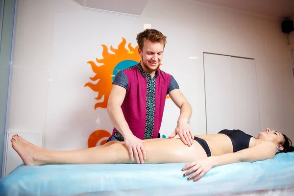 El concepto de masaje y salud. Un masajista masculino hace drenaje linfático y masaje para tonificar los músculos de una chica en ropa interior negra sobre una mesa en una sala fisiológica . — Foto de Stock