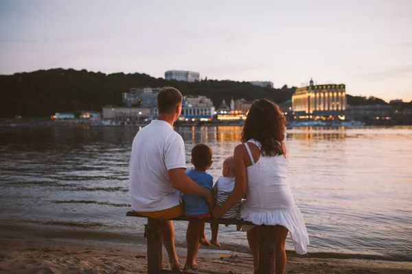 Concepto vacaciones en la playa familiar. Familia de cuatro personas mamá, papá y hermanos niños niños sentados en la orilla del río con la espalda en un banco de madera al atardecer en verano —  Fotos de Stock