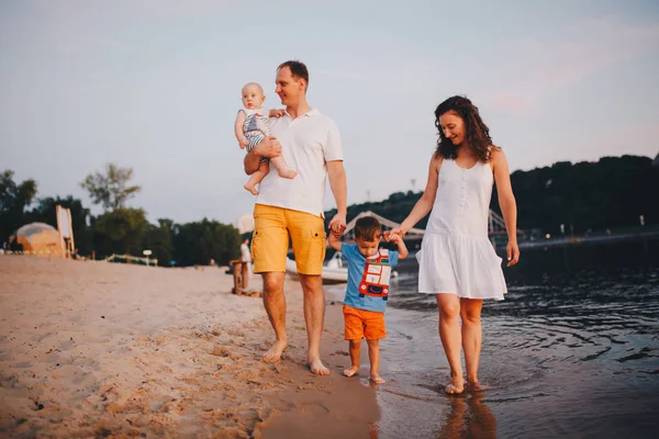 Concepto vacaciones en la playa familiar. Familia de cuatro personas mamá, papá y hermanos niños niños caminando, quieren descalzos en la orilla del río por la mano al atardecer en el verano —  Fotos de Stock