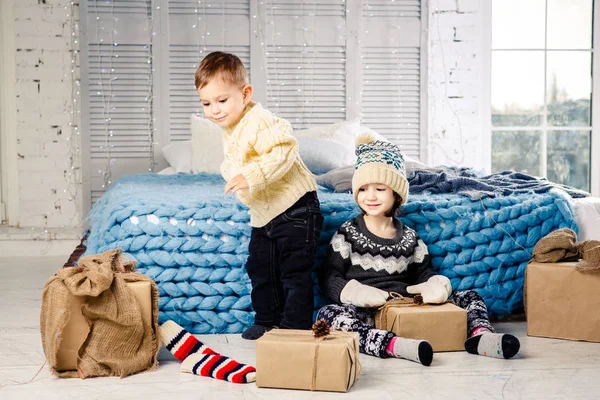 Los niños hermano y hermana se sientan en el suelo en el dormitorio cerca de la cama con regalos en el fondo de la decoración de Navidad en un día soleado. Vestido con una cálida prenda de punto de lana gorra y manoplas de colores — Foto de Stock