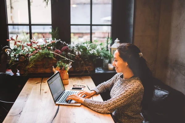 Mooie jonge brunette vrouw met behulp van een laptop bij coffeeshop op een houten tafel in de buurt van venster tekst typen op een toetsenbord. In de winter, met het licht van de lamp, is ze gekleed in een warme grijze trui — Stockfoto