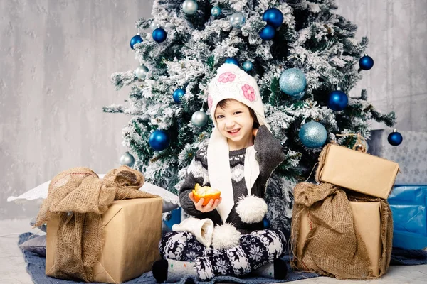 Niña de cinco años sentada en el suelo cerca del árbol de Navidad decorado con juguetes, pelotas. En las manos tiene una mandarina fruta naranja, naranja. vestido con una carpeta divertida y un suéter gris — Foto de Stock