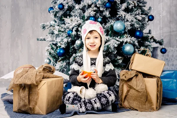 Niña de cinco años sentada en el suelo cerca del árbol de Navidad decorado con juguetes, pelotas. En las manos tiene una mandarina fruta naranja, naranja. vestido con una carpeta divertida y un suéter gris — Foto de Stock