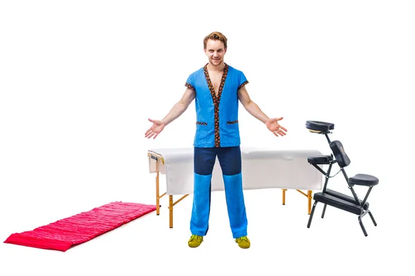 Masseur masculin en uniforme bleu se tient avec geste de saluer les mains sur les côtés avec le sourire à côté de l'équipement pour table de massage, chaise pliante, tapis rouge sur fond blanc. concept de beauté et de santé — Photo