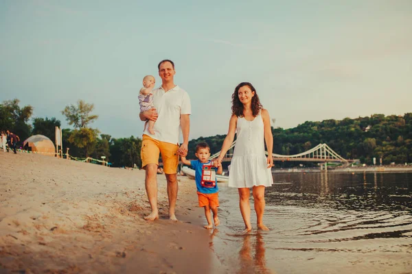Concepto vacaciones en la playa familiar. Familia de cuatro personas mamá, papá y hermanos niños niños caminando, quieren descalzos en la orilla del río por la mano al atardecer en el verano —  Fotos de Stock