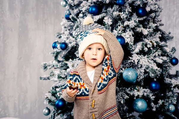 Pequeño niño divertido en tocado y suéter de lana corporal come dulces de chocolate en el fondo de un árbol de Navidad en la víspera de la Navidad home.He comió y untó sus mejillas, sosteniendo su sombrero con una mano — Foto de Stock