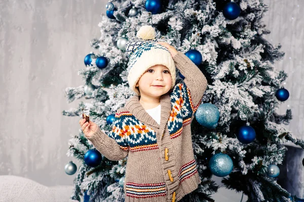 Μικρό αστείο αγόρι στο σώμα και κόμμωση μάλλινα πουλόβερ τρώει σοκολάτα γλυκά στο φόντο ενός χριστουγεννιάτικου δέντρου την παραμονή των Χριστουγέννων σπίτι. Έτρωγε και αλείψει τα μάγουλά του, κρατώντας το καπέλο με το ένα χέρι — Φωτογραφία Αρχείου