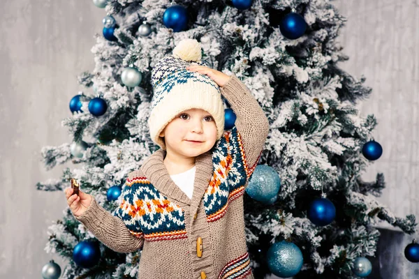 Pequeño niño divertido en tocado y suéter de lana corporal come dulces de chocolate en el fondo de un árbol de Navidad en la víspera de la Navidad home.He comió y untó sus mejillas, sosteniendo su sombrero con una mano — Foto de Stock