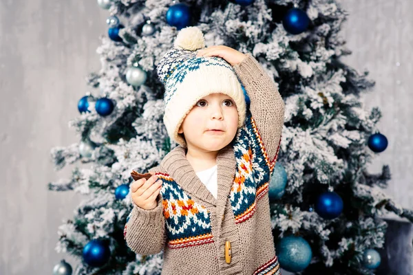 Pequeño niño divertido en tocado y suéter de lana corporal come dulces de chocolate en el fondo de un árbol de Navidad en la víspera de la Navidad home.He comió y untó sus mejillas, sosteniendo su sombrero con una mano — Foto de Stock