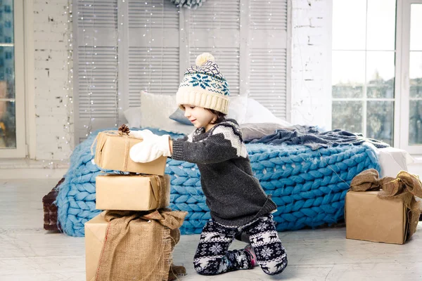 Pequeña niña está doblando una montaña de cajas de regalo en casa, cerca de la cama. El interior está decorado con decoración navideña. Luz solar brillante durante el día desde la ventana. Tema Año Nuevo — Foto de Stock