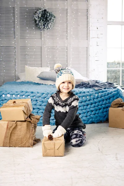 Pequena menina está dobrando uma montanha de caixas de presente em casa, perto da cama. O interior está decorado com decoração de Natal. Luz solar brilhante diurna da janela. Ano Novo tema — Fotografia de Stock