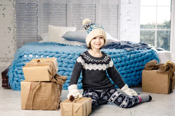 Pequeña niña está doblando una montaña de cajas de regalo en casa, cerca de la cama. El interior está decorado con decoración navideña. Luz solar brillante durante el día desde la ventana. Tema Año Nuevo — Foto de Stock