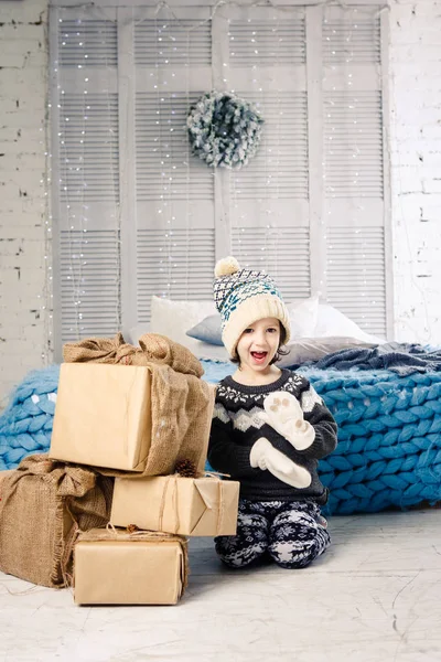 Pequeña niña está doblando una montaña de cajas de regalo en casa, cerca de la cama. El interior está decorado con decoración navideña. Luz solar brillante durante el día desde la ventana. Tema Año Nuevo — Foto de Stock