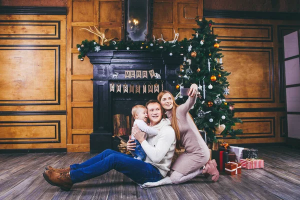 Tema navideño. joven familia con niño rubio de un año se sienta en el suelo de madera contra el fondo de un árbol de Navidad con regalos y hace selfie, autorretrato en la cámara frontal de un teléfono de plata — Foto de Stock