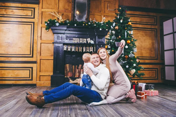 Різдвяна тема. молода сім'я з блондинкою одного року сидить на дерев'яній підлозі на фоні ялинки з подарунками і робить селфі, автопортрет на передній камері срібного телефону — стокове фото