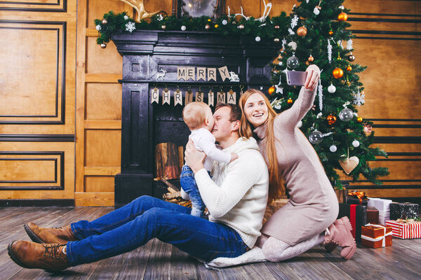 Christmas theme. young family with blond boy of one year sits on wooden floor against background of a Christmas tree with gifts and makes selfie, self-portrait on the front camera of a silver phone
