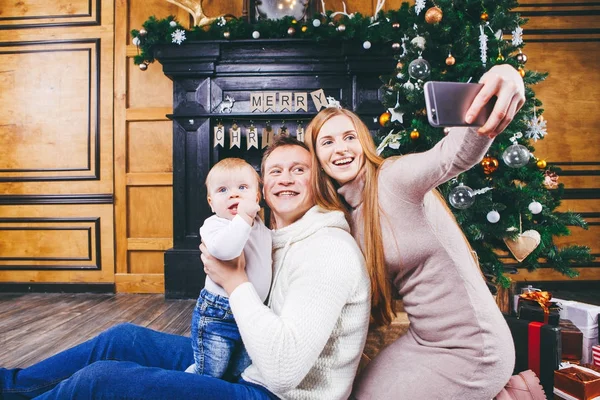 Tema navideño. joven familia con niño rubio de un año se sienta en el suelo de madera contra el fondo de un árbol de Navidad con regalos y hace selfie, autorretrato en la cámara frontal de un teléfono de plata — Foto de Stock