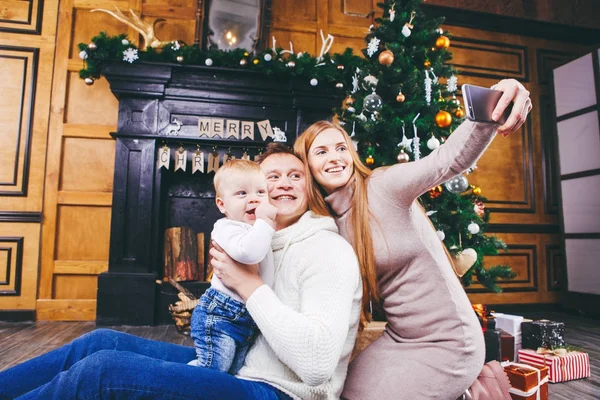 Tema navideño. joven familia con niño rubio de un año se sienta en el suelo de madera contra el fondo de un árbol de Navidad con regalos y hace selfie, autorretrato en la cámara frontal de un teléfono de plata — Foto de Stock