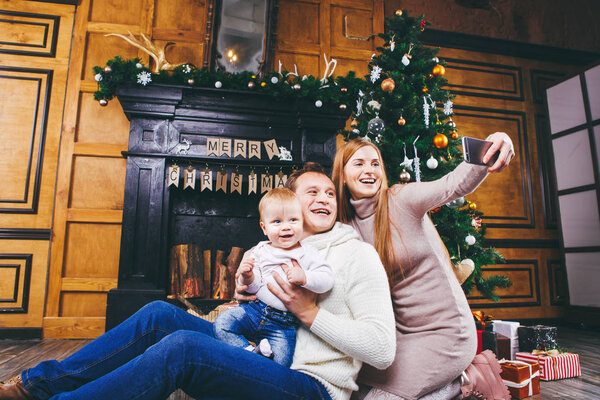 Christmas theme. young family with blond boy of one year sits on wooden floor against background of a Christmas tree with gifts and makes selfie, self-portrait on the front camera of a silver phone