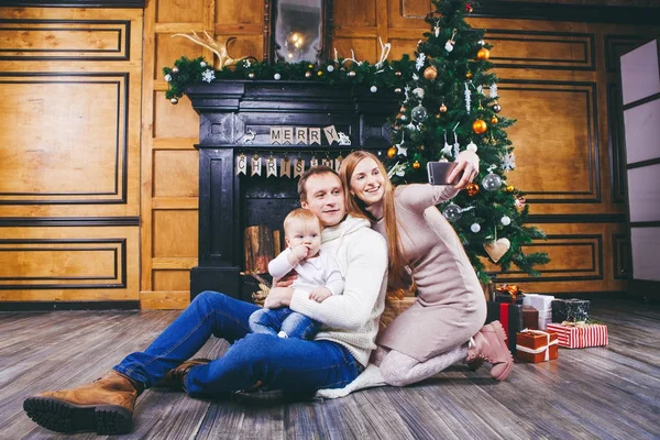 Christmas theme. young family with blond boy of one year sits on wooden floor against background of a Christmas tree with gifts and makes selfie, self-portrait on the front camera of a silver phone — Stock Photo, Image