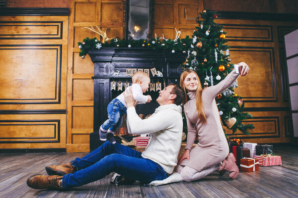 Christmas theme. Father throws the son of blond one year old, sit on a wooden tree in front of Christmas tree with gifts, mom makes selfie, self-portrait on the front camera phone of a silver color