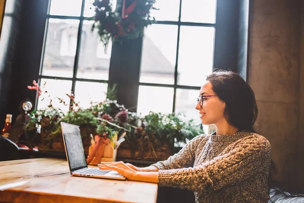 Mooie brunette zakelijke vrouw zakenvrouw maakt gebruik van een laptop, kiest, wordt tekst afgedrukt op het toetsenbord. In een koffiehuis, zitten aan een tafel in de buurt van een venster. Het interieur is versierd met kerst decor — Stockfoto