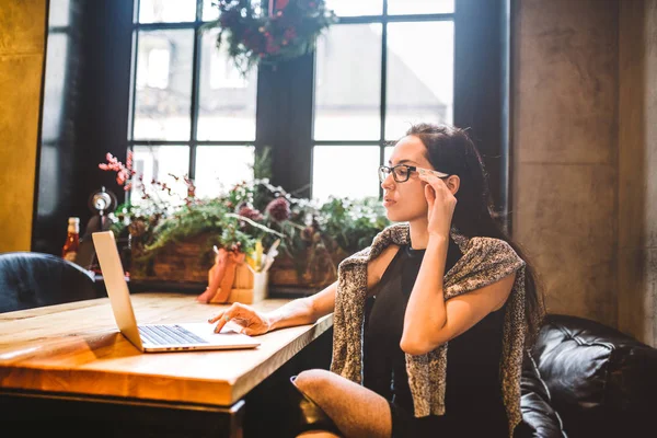 Όμορφη νεαρή μελαχρινή επιχειρήσεων γυναίκα που φοράει γυαλιά και πουλόβερ χρησιμοποιεί φορητό υπολογιστή για να εκτυπώσετε κείμενο στο πληκτρολόγιο. Στο σπίτι καφέ, κάθονται στο τραπέζι κοντά στο παράθυρο. Το εσωτερικό είναι διακοσμημένο με Χριστουγεννιάτικη διακόσμηση — Φωτογραφία Αρχείου