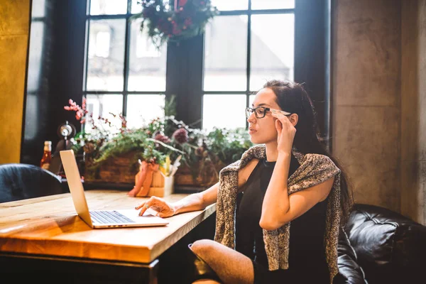 Mooie jonge brunette zakenvrouw dragen van bril en trui maakt gebruik van laptop tekst afdrukken op toetsenbord. In koffiehuis, zittend aan tafel in de buurt van venster. Het interieur is versierd met kerst decor — Stockfoto