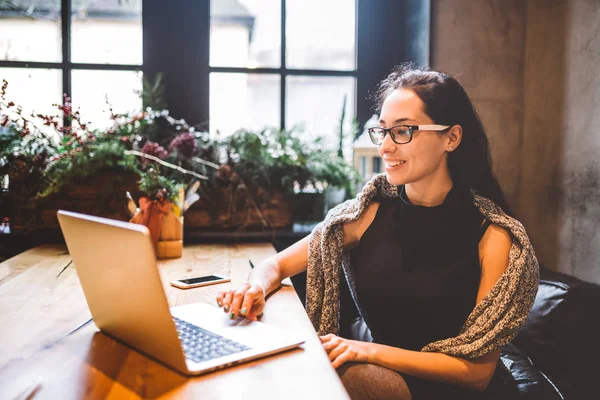 Mooie jonge brunette zakenvrouw dragen van bril en trui maakt gebruik van laptop tekst afdrukken op toetsenbord. In koffiehuis, zittend aan tafel in de buurt van venster. Het interieur is versierd met kerst decor — Stockfoto